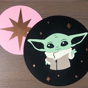 Pode incluir: Um c&iacute;rculo preto com um personagem verde da franquia Star Wars, Baby Yoda, com um casaco marrom e uma gola branca. O personagem est&aacute; cercado por pequenas estrelas douradas em um fundo preto. Um c&iacute;rculo rosa com uma estrela dourada est&aacute; no fundo.