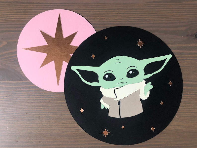 Pode incluir: Um c&iacute;rculo preto com um personagem verde da franquia Star Wars, Baby Yoda, com um casaco marrom e uma gola branca. O personagem est&aacute; cercado por pequenas estrelas douradas em um fundo preto. Um c&iacute;rculo rosa com uma estrela dourada est&aacute; no fundo.
