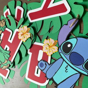 May include: A colorful paper banner with the letters "Happy" and a blue Stitch character from the Disney movie Lilo & Stitch. The banner also features green leaves, pink flowers, and a brown string.