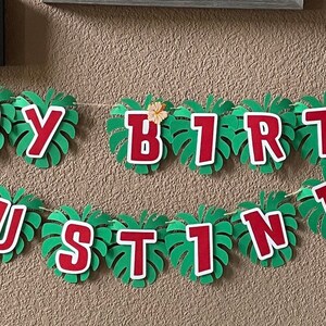 May include: A green and red banner with the words "Happy Birthday Justine" and a blue Stitch character. The banner is decorated with green leaves.