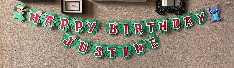 May include: A green and red banner with the words "Happy Birthday Justine" and a blue Stitch character. The banner is decorated with green leaves.