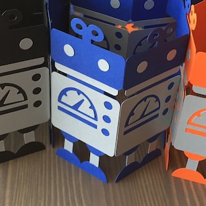May include: Three papercraft robots, one black, one blue, and one orange, with silver accents. Each robot has a speedometer on its chest and a pair of antennas on its head.