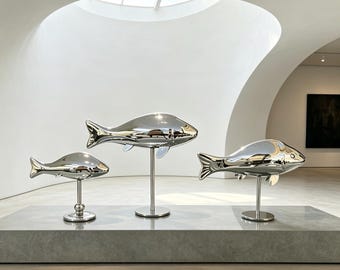 May include: Three polished silver-toned metal fish sculptures mounted on individual vertical stands. Each fish features a smooth, reflective surface and is positioned at a different height, creating a tiered arrangement of aquatic figures.