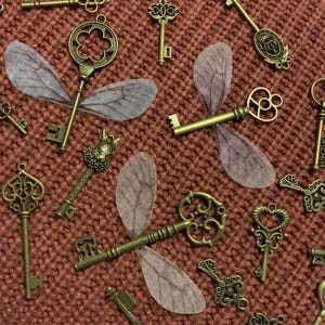 May include: A collection of antique-style bronze-toned metal keys in various intricate designs. Several keys feature translucent, veined, wing-like attachments extending from their shafts, resembling insects. The keys vary in size and shape.