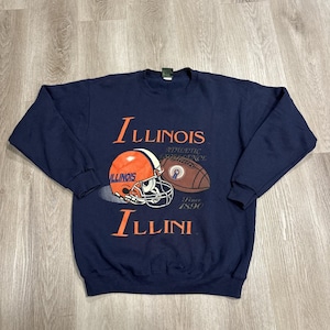 Puede incluir: Una sudadera azul marino de manga larga con el texto Illinois e Illini en naranja. El gráfico incluye un casco de fútbol americano naranja y blanco, un balón de fútbol americano marrón y el texto Athletic Excellence Since 1890 en blanco.