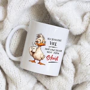 Könnte beinhalten: Eine weiße Keramiktasse mit einer Illustration einer Ente mit zerzaustem Gefieder, die eine kleine Tasse hält. Der Text lautet: ICH SCHLUCKE VIEL ABER GANZ SICHER NICHT JEDEN Scheiß. Die Ente hat orangefarbene Füße und einen Schnabel.