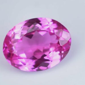 Può includere: Una gemma dal taglio ovale di un colore rosa intenso con una superficie sfaccettata che riflette la luce. La pietra presenta una disposizione simmetrica di sfaccettature triangolari e a forma di diamante su tutta la superficie superiore.