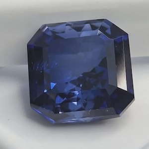 Può includere: Una pietra preziosa di colore blu intenso con taglio quadrato e superficie sfaccettata che crea complessi riflessi di luce. La pietra presenta una ricca tonalità indaco satura con bordi netti e puliti e una forma geometrica simmetrica.