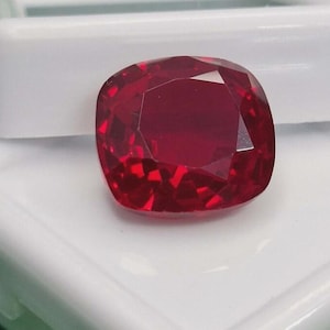 Peut inclure: Une pierre précieuse taille coussin d'une couleur rouge profonde et vibrante, dotée de multiples facettes qui reflètent la lumière. La pierre est posée sur une surface blanche lisse et rectangulaire, révélant sa transparence et sa forme polie.
