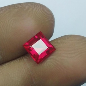 May include: A vibrant, square-cut red gemstone with a faceted surface that reflects light. The stone features sharp, clean edges and a deep, saturated crimson hue, showcasing a clear, geometric structure with a polished, reflective finish.