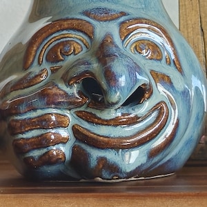 May include: A ceramic vessel featuring a sculpted face with a prominent nose, wide eyes, and a smiling mouth. The piece is finished in a glossy glaze with a blend of light blue and deep brown tones, highlighting the textured facial features.