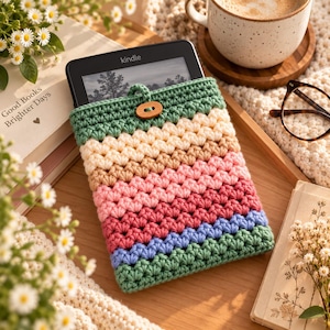May include: A crochet e-reader sleeve featuring horizontal stripes in sage green, cream, tan, light pink, dark pink, and periwinkle blue. The sleeve has a sage green top flap secured by a small, oval-shaped wooden button closure.