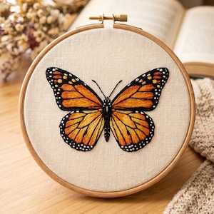 May include: A circular wooden embroidery hoop containing a piece of off-white fabric. Centered on the fabric is a detailed hand-stitched monarch butterfly featuring vibrant orange and black thread with small white accents on the wing edges.