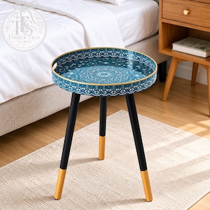 May include: A round side table featuring a teal tray top with an intricate white mandala pattern and a gold rim. The table stands on three black legs, each finished with a gold-colored tip at the base, providing a stable structure for the tray.