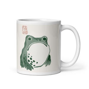 May include: A white ceramic mug featuring a green frog illustration with a white belly and dark green spots. Above the frog is a small red square stamp with Asian characters. The mug has a smooth finish, a curved handle, and a cylindrical shape.