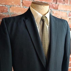 May include: A dark navy blue blazer worn over a cream-colored button-down shirt and a patterned olive green tie. The blazer features notched lapels and a structured fit, displayed on a mannequin torso against a textured red brick wall background.