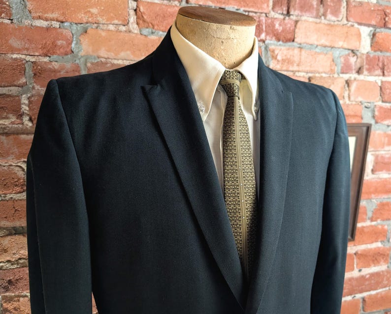 May include: A dark navy blue blazer worn over a cream-colored button-down shirt and a patterned olive green tie. The blazer features notched lapels and a structured fit, displayed on a mannequin torso against a textured red brick wall background.