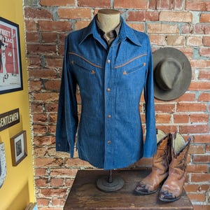 K&ouml;nnte beinhalten: Ein blaues Jeanshemd mit beigen N&auml;hten und Kn&ouml;pfen, auf einer Schaufensterpuppe pr&auml;sentiert. Das Hemd hat zwei Brusttaschen und einen klassischen Kragen. Ein Paar braune Cowboy-Stiefel und ein Hut sind in der N&auml;he.