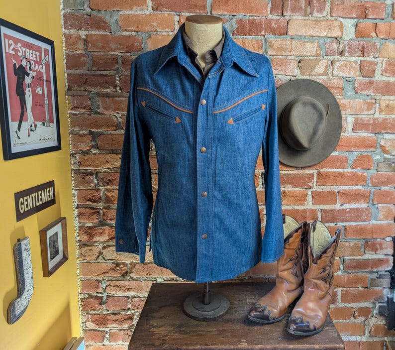 K&ouml;nnte beinhalten: Ein blaues Jeanshemd mit beigen N&auml;hten und Kn&ouml;pfen, auf einer Schaufensterpuppe pr&auml;sentiert. Das Hemd hat zwei Brusttaschen und einen klassischen Kragen. Ein Paar braune Cowboy-Stiefel und ein Hut sind in der N&auml;he.