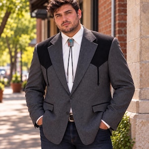 May include: A charcoal grey blazer featuring black shoulder yokes and black pocket trim, worn over a crisp white collared shirt. A black bolo tie with a central turquoise and red stone inlay is fastened at the collar, paired with dark blue denim jeans.