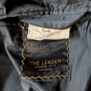 May include: A dark navy blue garment interior featuring a black rectangular fabric label with gold stitching. The label reads Eagle Clothes, The Leader, Kansas City, Kansas. A white union label is stitched above with Amalgamated Clothing text.