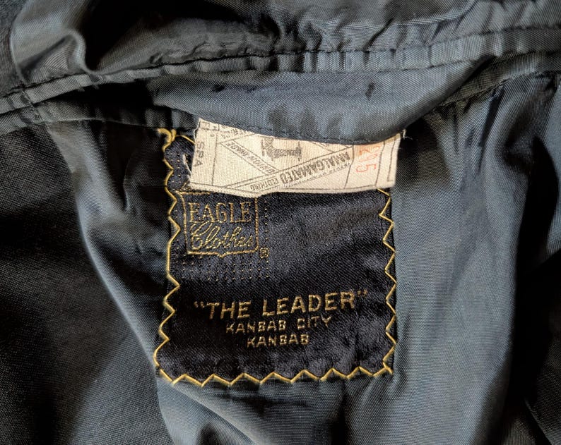 May include: A dark navy blue garment interior featuring a black rectangular fabric label with gold stitching. The label reads Eagle Clothes, The Leader, Kansas City, Kansas. A white union label is stitched above with Amalgamated Clothing text.