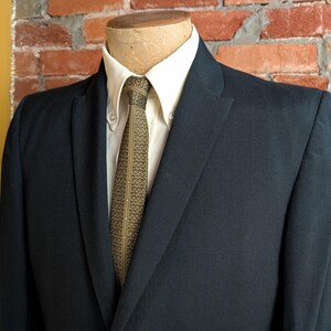 May include: A dark navy blue suit jacket worn over a crisp white button-down shirt. The ensemble is completed with a textured olive green necktie featuring a repeating geometric pattern and a thin vertical line of small white dots down the center.