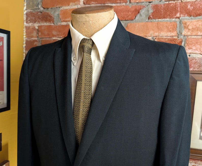 May include: A dark navy blue suit jacket worn over a crisp white button-down shirt. The ensemble is completed with a textured olive green necktie featuring a repeating geometric pattern and a thin vertical line of small white dots down the center.