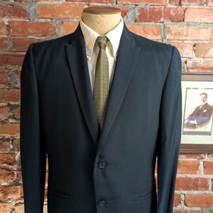 May include: A dark charcoal suit jacket with a two-button closure, worn over a cream-colored button-down shirt and a textured olive green necktie with a subtle geometric pattern. The jacket features notched lapels and two front flap pockets.
