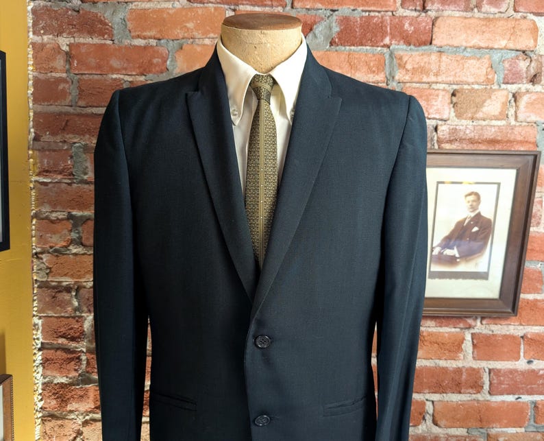 May include: A dark charcoal suit jacket with a two-button closure, worn over a cream-colored button-down shirt and a textured olive green necktie with a subtle geometric pattern. The jacket features notched lapels and two front flap pockets.