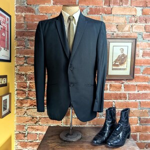 May include: A black two-button blazer worn over a light-colored collared shirt and a textured olive green necktie. The blazer features a notched lapel, two front flap pockets, and long sleeves, displayed on a mannequin stand with black boots.