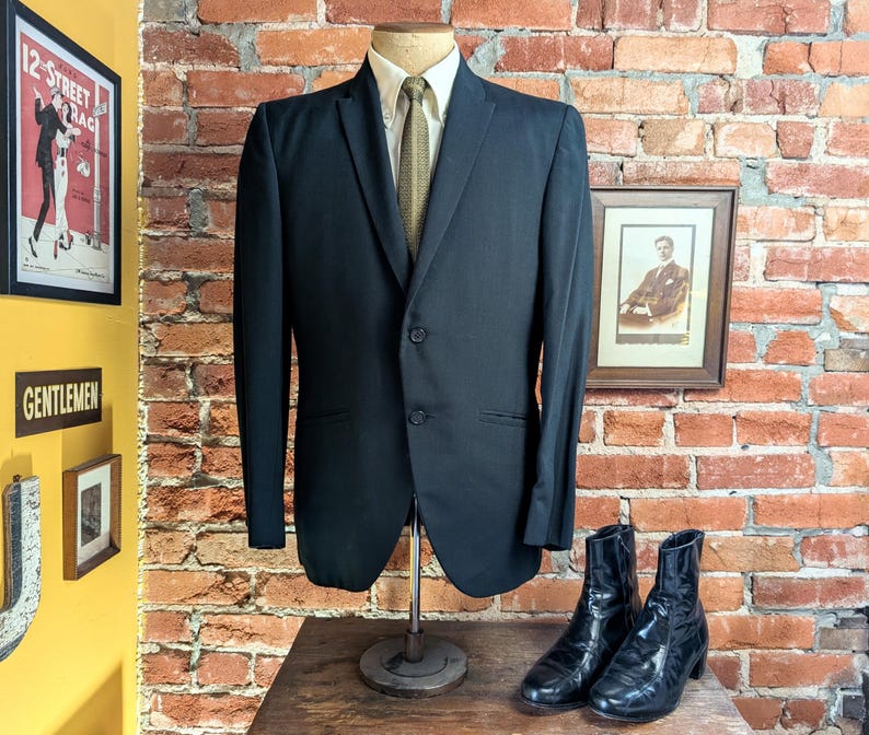 May include: A black two-button blazer worn over a light-colored collared shirt and a textured olive green necktie. The blazer features a notched lapel, two front flap pockets, and long sleeves, displayed on a mannequin stand with black boots.