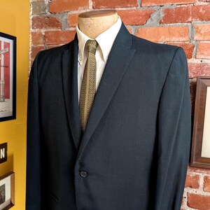 May include: A dark navy blue blazer worn over a crisp white button-down shirt and a textured olive green necktie with a subtle geometric pattern. The blazer features a single-button closure, notched lapels, and structured shoulders on a mannequin.