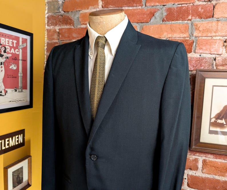 May include: A dark navy blue blazer worn over a crisp white button-down shirt and a textured olive green necktie with a subtle geometric pattern. The blazer features a single-button closure, notched lapels, and structured shoulders on a mannequin.