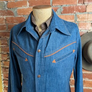 K&ouml;nnte beinhalten: Eine blaue Jeansjacke mit orangefarbenen N&auml;hten und Akzenten. Die Jacke hat ein klassisches Design mit Kragen und Knopfverschluss. Die Jacken wird auf einer Schaufensterpuppe pr&auml;sentiert.