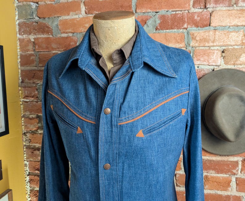 K&ouml;nnte beinhalten: Eine blaue Jeansjacke mit orangefarbenen N&auml;hten und Akzenten. Die Jacke hat ein klassisches Design mit Kragen und Knopfverschluss. Die Jacken wird auf einer Schaufensterpuppe pr&auml;sentiert.