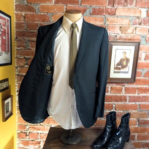 May include: A dark charcoal blazer worn over a crisp white button-down shirt and a textured olive green necktie. The blazer is displayed on a mannequin, showing a black interior lining with a small rectangular label featuring gold text.
