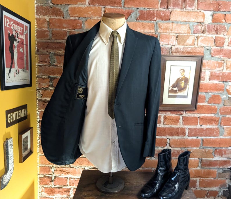 May include: A dark charcoal blazer worn over a crisp white button-down shirt and a textured olive green necktie. The blazer is displayed on a mannequin, showing a black interior lining with a small rectangular label featuring gold text.