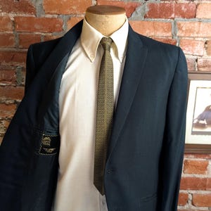 May include: A dark navy blue blazer worn over a cream-colored button-down shirt and a patterned olive green tie. The interior lining of the blazer features a black label with the text "THE LEADER" and "MADE IN USA" visible on the left side.