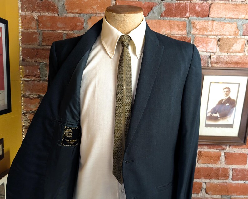 May include: A dark navy blue blazer worn over a cream-colored button-down shirt and a patterned olive green tie. The interior lining of the blazer features a black label with the text "THE LEADER" and "MADE IN USA" visible on the left side.