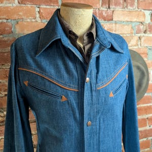 K&ouml;nnte beinhalten: Eine blaue Jeansjacke im Western-Stil. Die Jacke hat braune Zierleisten und passende Kn&ouml;pfe. Die Jacke wird auf einer Schaufensterpuppe vor einer Backsteinmauer pr&auml;sentiert. Der Kragen ist klassisch.