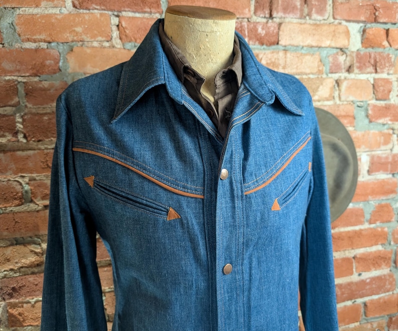 K&ouml;nnte beinhalten: Eine blaue Jeansjacke im Western-Stil. Die Jacke hat braune Zierleisten und passende Kn&ouml;pfe. Die Jacke wird auf einer Schaufensterpuppe vor einer Backsteinmauer pr&auml;sentiert. Der Kragen ist klassisch.