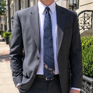 May include: A charcoal gray suit jacket worn over a crisp white button-down shirt and a dark navy blue necktie. The necktie features a subtle pattern of small dots and a detailed embroidered design of horses near the bottom tip of the fabric.