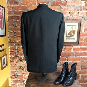 May include: A solid black blazer shown from the back, featuring a structured collar, long sleeves, and a single center vent at the bottom hem. The garment is displayed on a mannequin torso, showcasing a smooth, uniform fabric texture throughout.