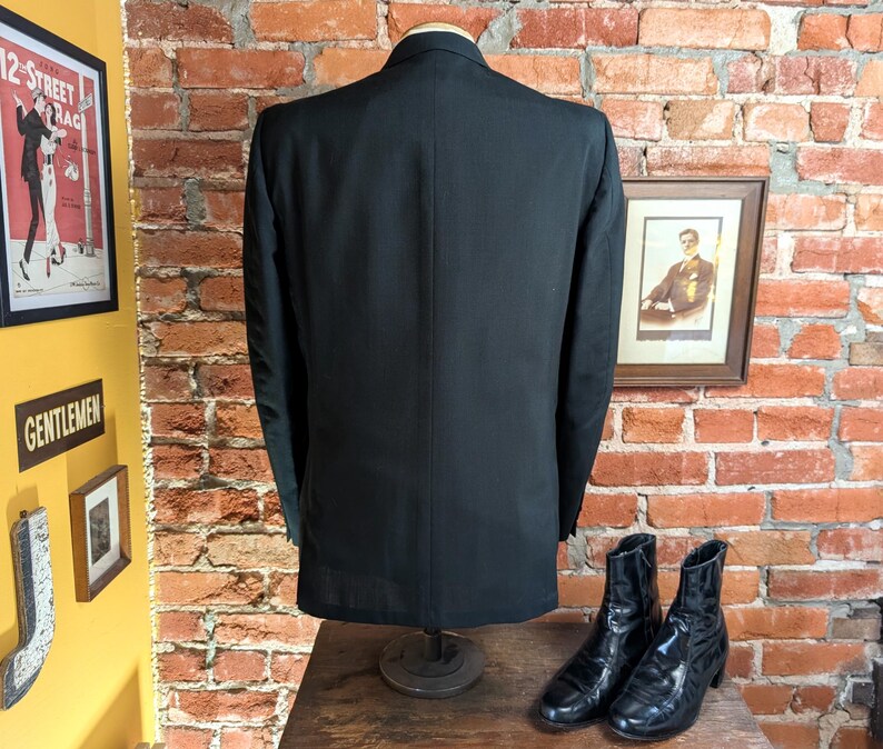 May include: A solid black blazer shown from the back, featuring a structured collar, long sleeves, and a single center vent at the bottom hem. The garment is displayed on a mannequin torso, showcasing a smooth, uniform fabric texture throughout.