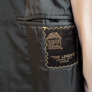May include: The interior of a black blazer featuring a black fabric label with gold embroidery. The label displays an eagle graphic, the text EAGLE Clothes, THE LEADER, and KANSAS CITY KANSAS. The blazer has a dark lining and a welt pocket.