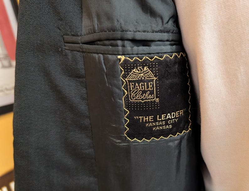 May include: The interior of a black blazer featuring a black fabric label with gold embroidery. The label displays an eagle graphic, the text EAGLE Clothes, THE LEADER, and KANSAS CITY KANSAS. The blazer has a dark lining and a welt pocket.
