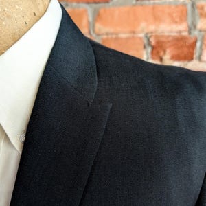 May include: A black blazer with a notched lapel worn over a crisp white collared shirt. The blazer features a structured shoulder and a smooth fabric texture, layered over a shirt with a visible button and a textured brown patterned tie.