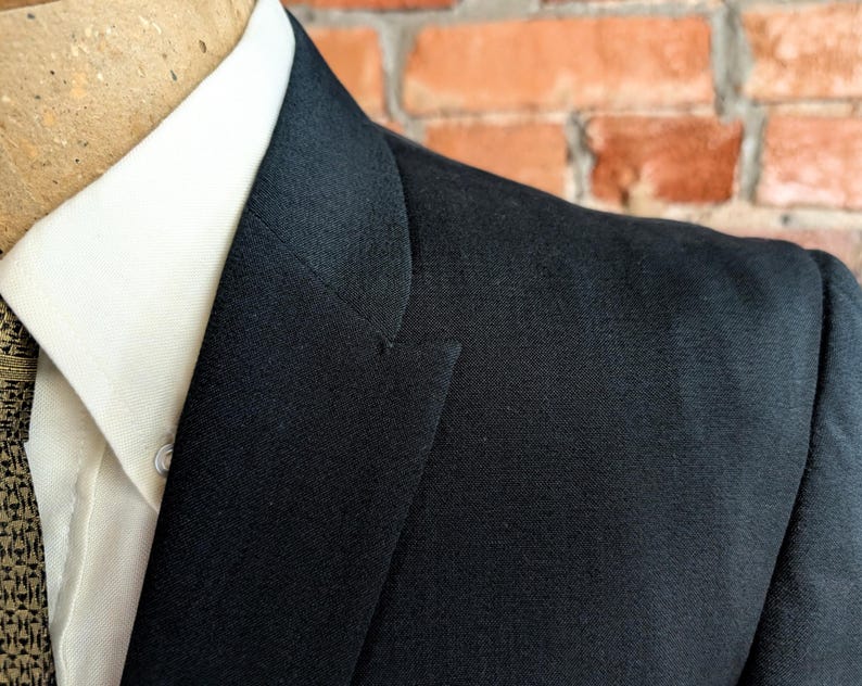 May include: A black blazer with a notched lapel worn over a crisp white collared shirt. The blazer features a structured shoulder and a smooth fabric texture, layered over a shirt with a visible button and a textured brown patterned tie.