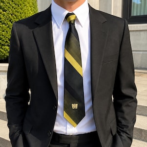 Puede incluir: Una chaqueta de traje negra sobre una camisa blanca de cuello rígido con una corbata de rayas diagonales negras y verde oliva. La corbata presenta un pequeño emblema bordado de color dorado cerca de la punta inferior, centrado en la tela.